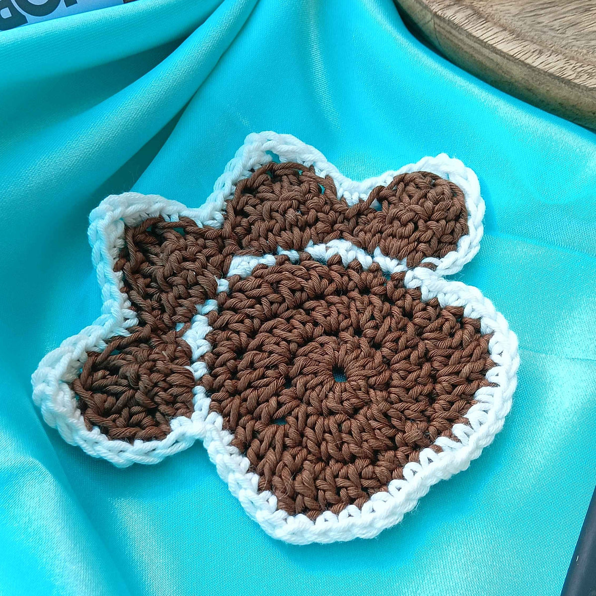 Crocheted brown and white paw shaped coaster on a turquoise fabric background